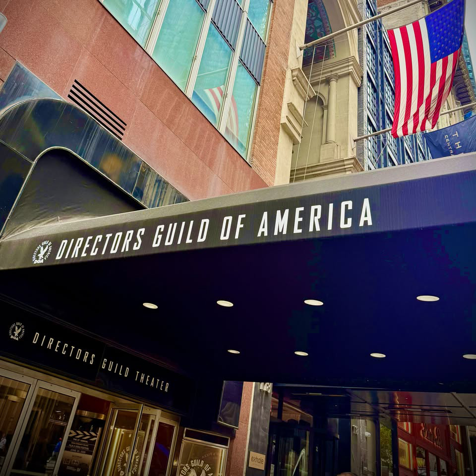 Exterior of the Directors Guild of America Theater, featuring its large marquee beneath an American flag and surrounding city buildings.